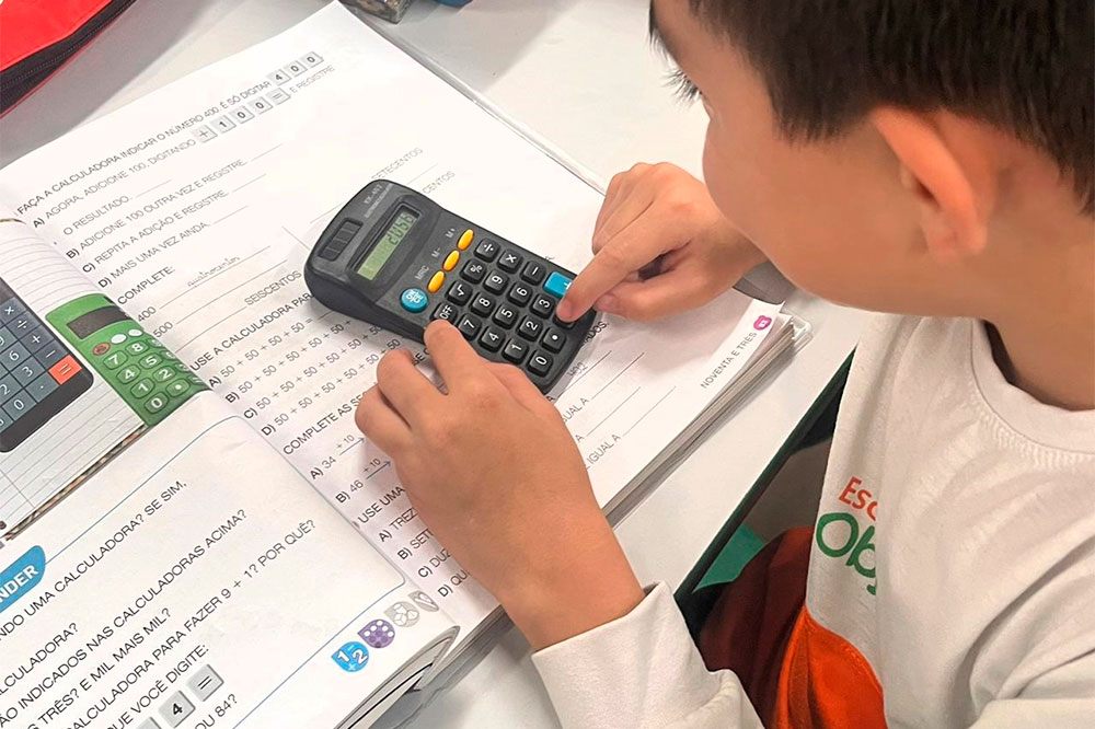 Criança utilizando uma calculadora enquanto realiza exercícios de matemática em um livro didático. O aluno, usando uniforme da Escola Objetiva, está concentrado, pressionando os botões da calculadora, que exibe um resultado no visor. O livro está aberto sobre a mesa e contém instruções e atividades relacionadas ao uso de calculadoras.